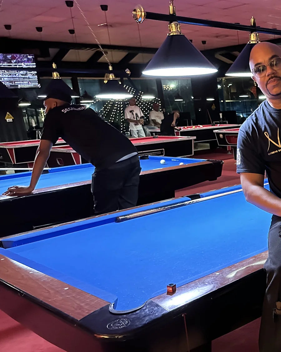 Adult man wearing a black Narcy wordmark T-shirt beside a pool table in a low-light editorial portrait